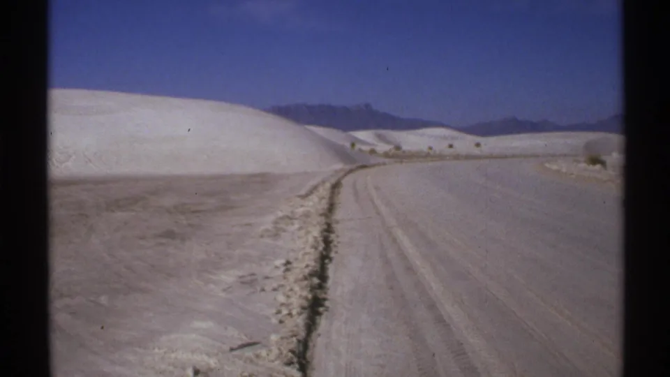 1972 New Mexico 8mm frame showing a dirt road curving through pale dune-like terrain toward distant mountains.