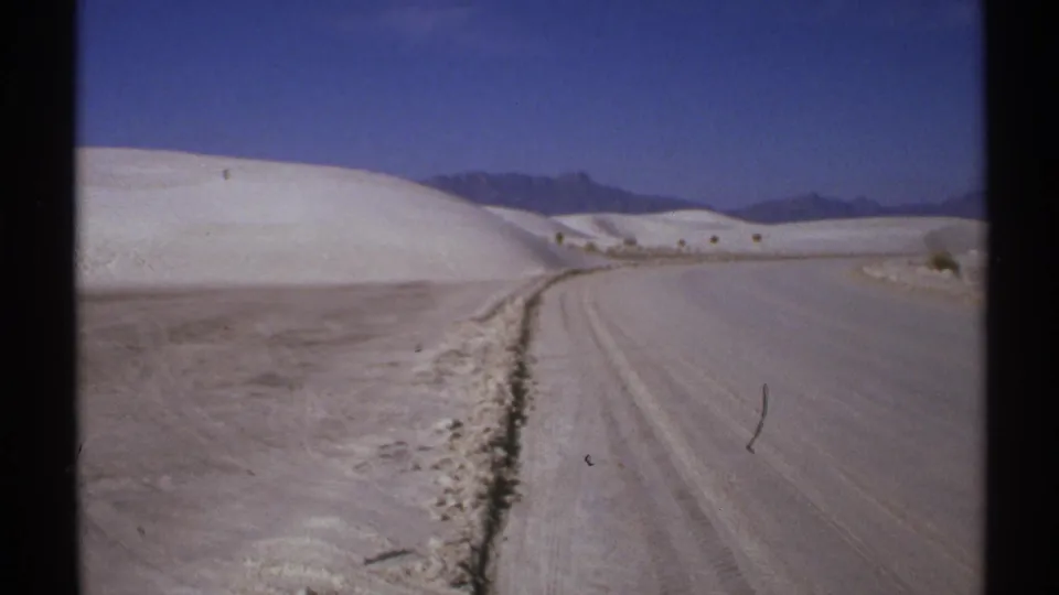 1972 New Mexico travel frame with a curving dirt road, pale dunes, sparse scrub, and distant mountain ridges.