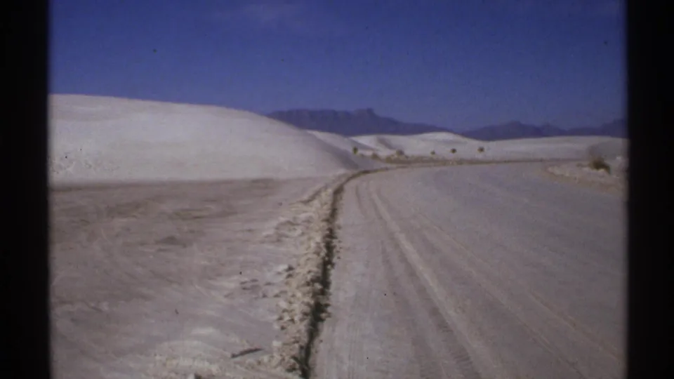 1972 New Mexico 8mm frame showing tire tracks on a dirt road running beside pale dunes and distant mountains.