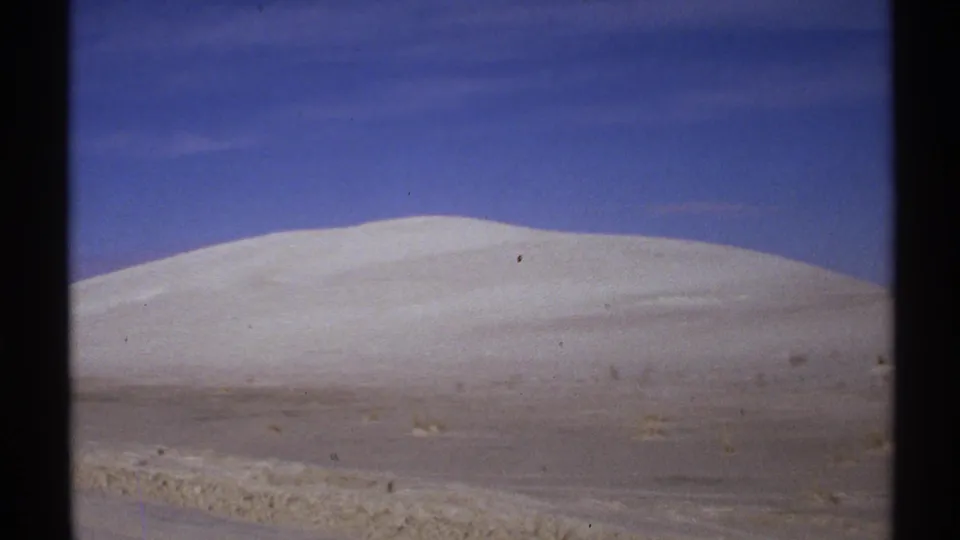 1972 New Mexico landscape frame showing a broad pale dune or snow-covered slope above sparse desert scrub.
