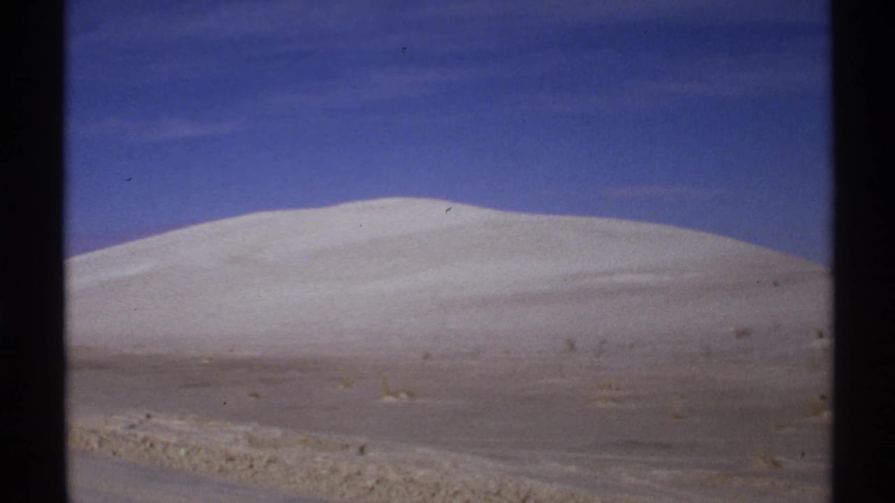 1972 New Mexico 8mm frame showing a broad pale dune, scrubby desert foreground, and open blue sky.