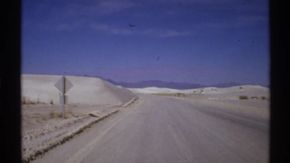 1972 New Mexico 8mm frame showing a paved road through pale dunes, a roadside sign, and distant low hills.