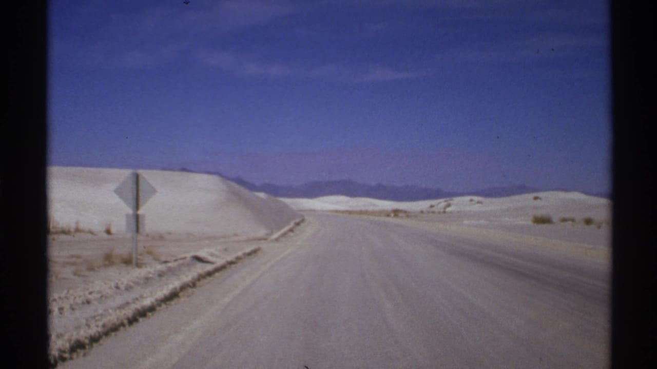 1972 8mm New Mexico road view with white sand dunes, a roadside sign, distant hills, and sparse desert vegetation.