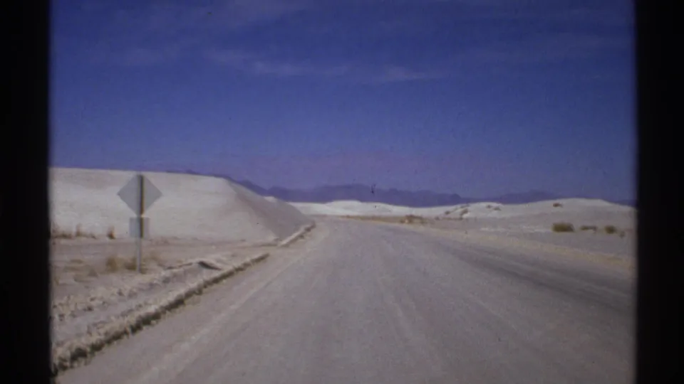 Vehicle-shot 8mm frame of a New Mexico road bordered by pale dunes, low mountains, and a small roadside sign.