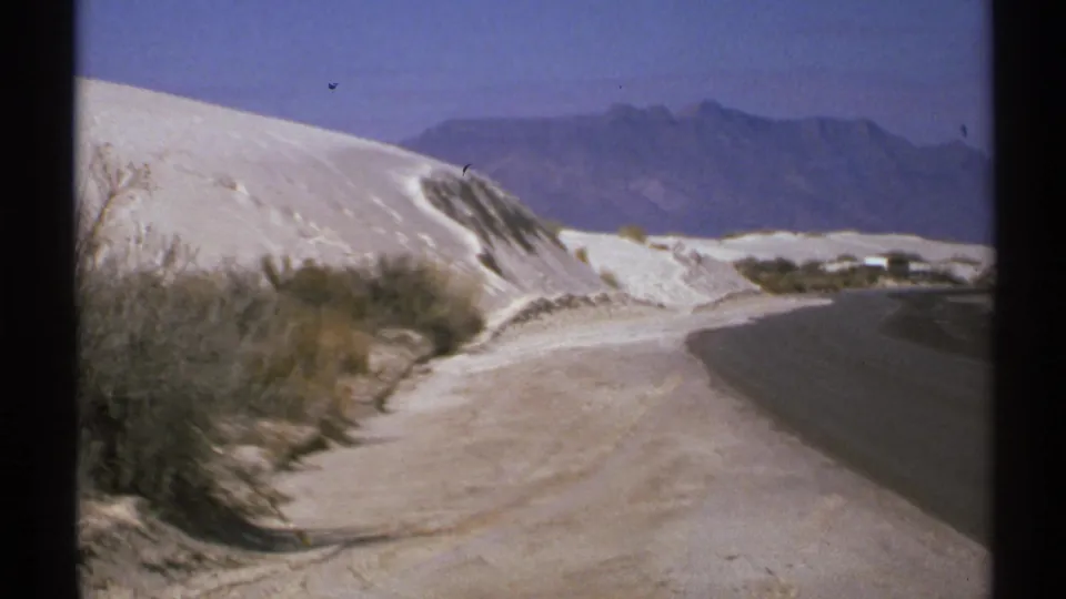 1972 New Mexico roadside view with white sand dunes, sparse desert brush, and distant mountains beyond the curve.