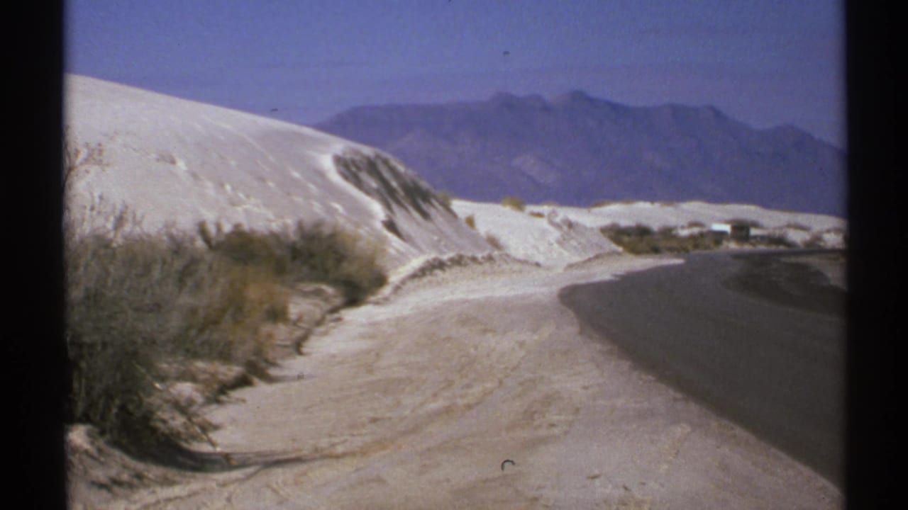 Curving road beside white sand dunes with shrubs and distant mountains.