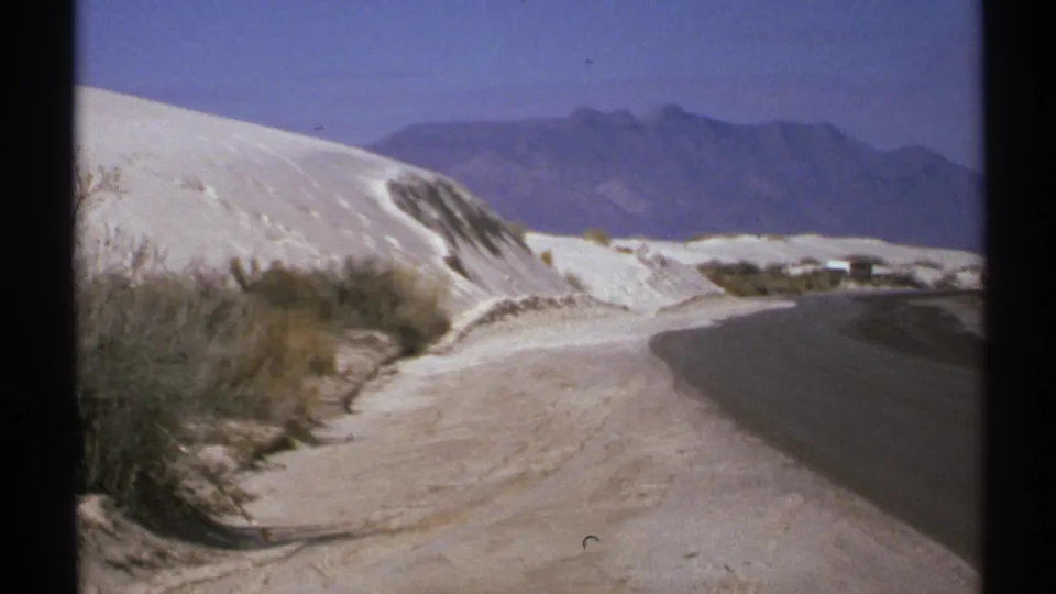 1972 New Mexico 8mm frame showing a paved road curving past white dunes, desert brush, and hazy mountains under blue sky.