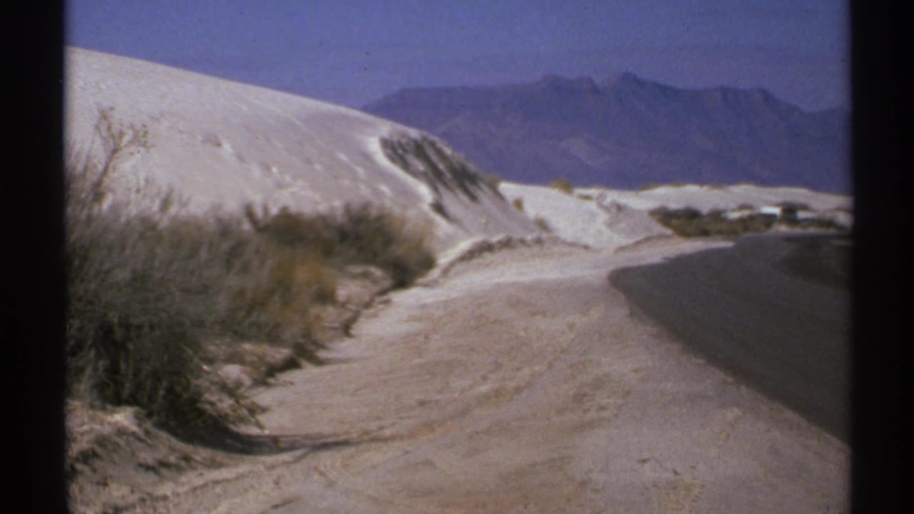 White dune slope and road curve with brush and mountains in the distance.