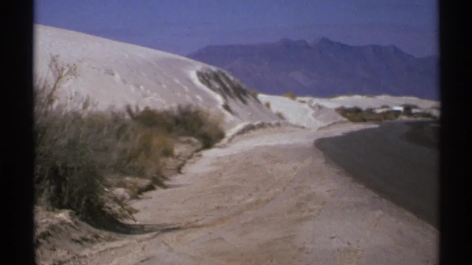 1972 New Mexico frame of a road skirting white sand dunes, with scrubby vegetation and blue mountain ridges on the horizon.