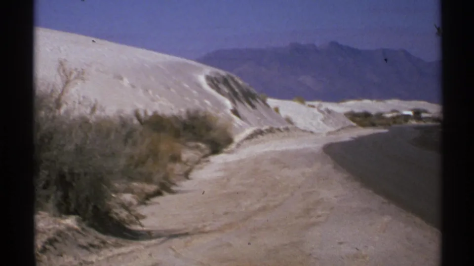 1972 New Mexico 8mm view of white roadside dunes, scrub vegetation, and a paved road leading toward distant mountains.