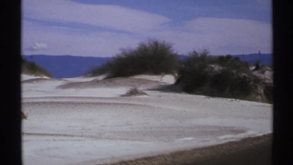 1972 8mm New Mexico view of pale desert dunes, sparse shrubs, distant mountains, and a bright blue sky.