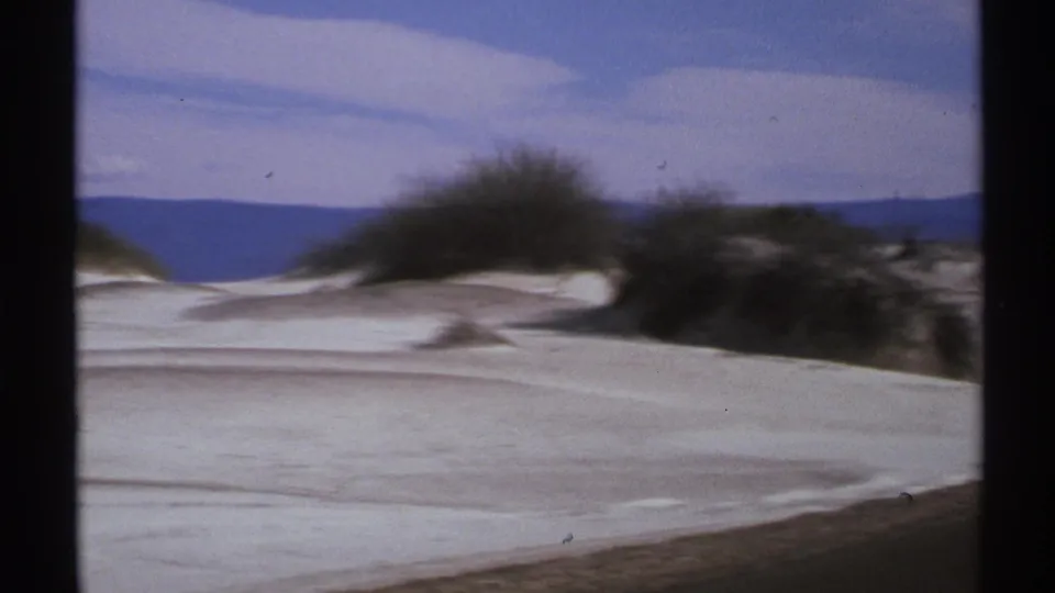 Soft 1972 8mm view of New Mexico dunes with dark desert brush and a blue mountain ridge in the distance.