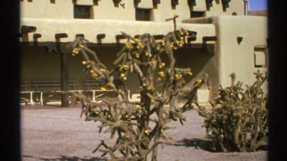 1972 8mm New Mexico frame showing an adobe-style building, shaded arcade, cactus, and desert planting.