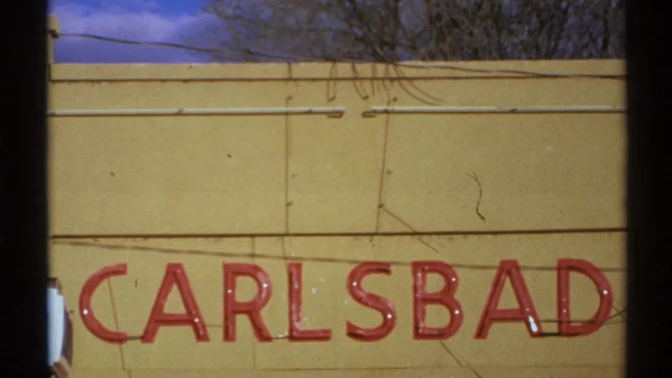 1972 New Mexico frame showing a red CARLSBAD sign on a yellow wall, with bare branches and blue sky above.