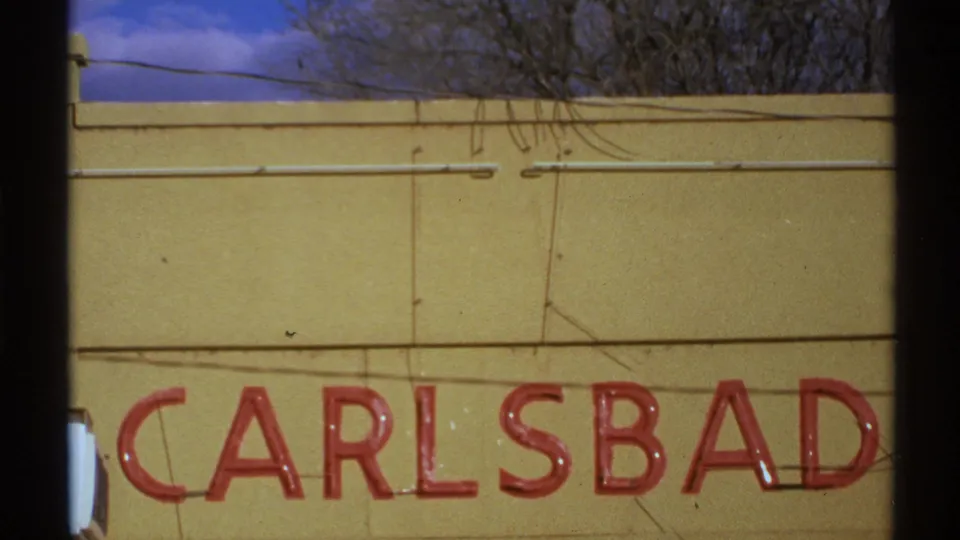 1972 New Mexico 8mm frame showing red CARLSBAD signage on a yellow facade, with wires, bare branches, and blue sky above.