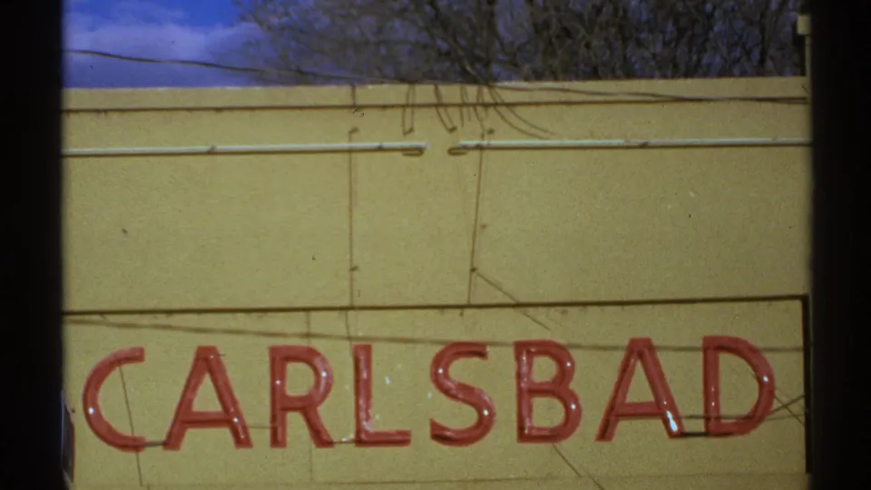 1972 New Mexico 8mm frame showing the CARLSBAD sign cropped low on a yellow exterior wall with overhead facade details.