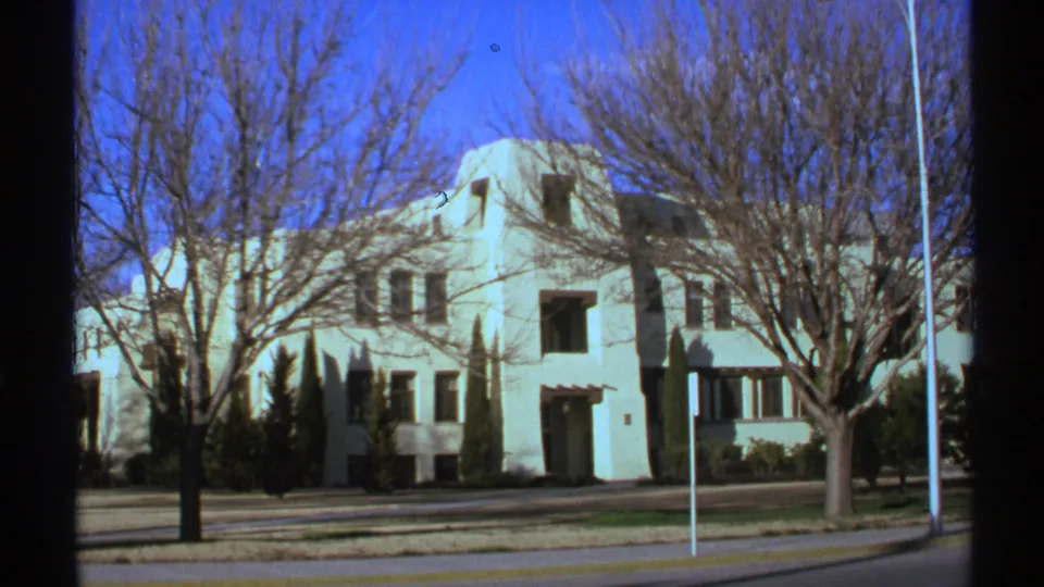 1972 New Mexico 8mm frame showing a white multi-story building across a street, with bare trees, flagpoles, and clear blue sky.