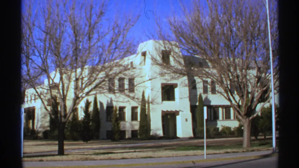 The same New Mexico building exterior appears in a steady 8mm view, partially obscured by bare tree branches.