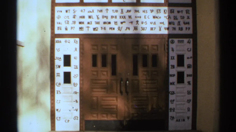 1972 New Mexico 8mm frame showing a wooden double door with glass panels beneath a projected grid of handwritten letters and symbols.
