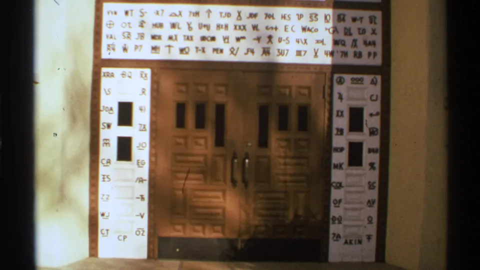 Final frame shows the same New Mexico home-movie view of a wooden double door framed by projected handwritten letters, numbers, and symbols.