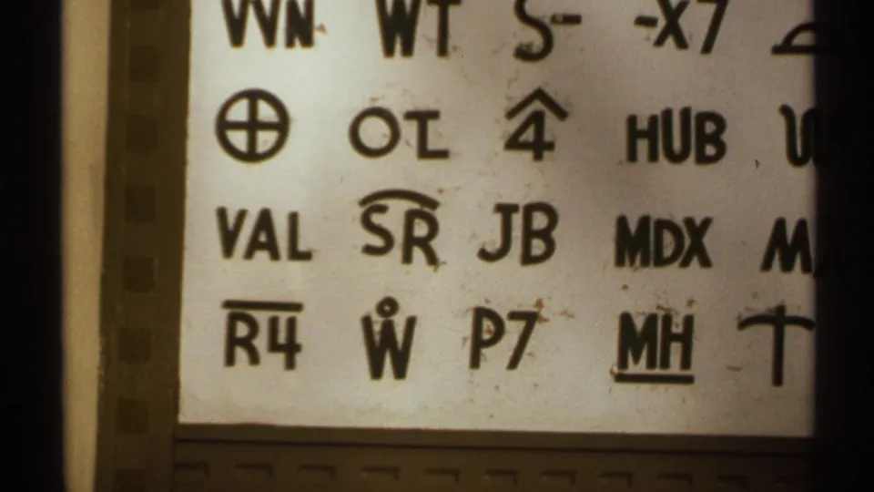 Close 8mm view of black alphanumeric codes and symbols on a white sign panel in the 1972 New Mexico clip.