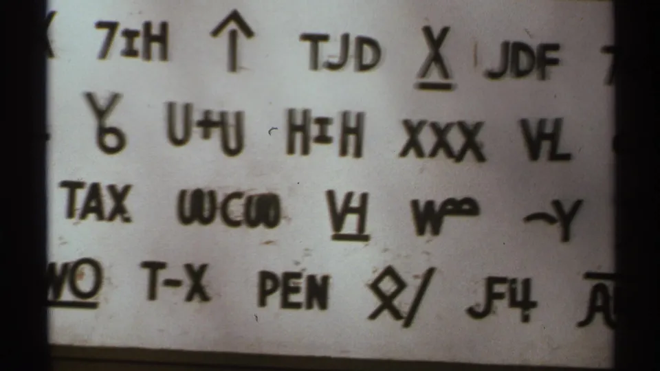 1972 8mm close-up of a white sheet filled with black handwritten codes, arrows, underlines, and block-like letter groups.