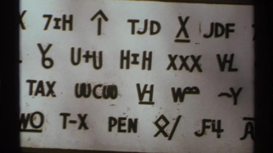 Close 8mm frame showing rows of coded-looking letters, numbers, arrows, and underlined marks on a white paper surface.