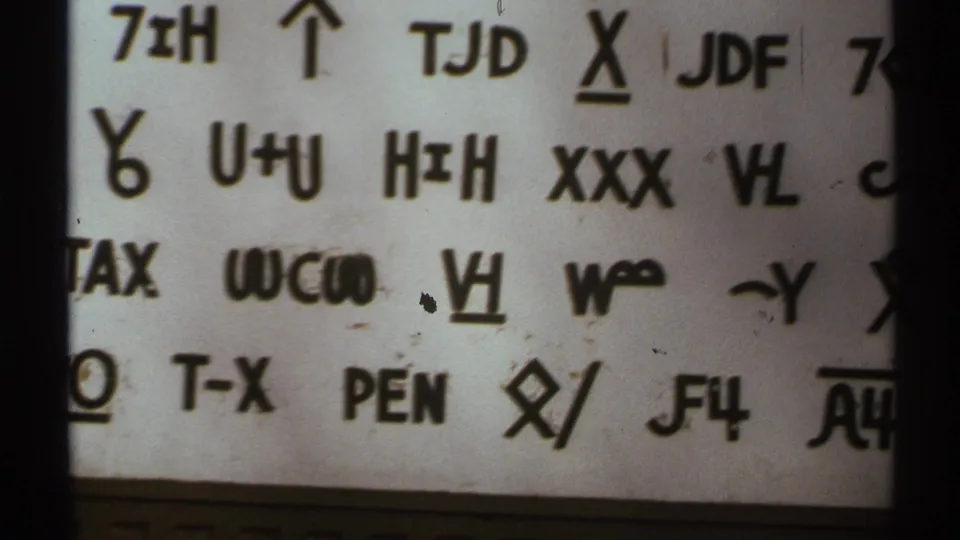 Soft 1972 8mm close-up of hand-drawn codes and symbols on white paper, framed by dark film edges and visible specks.