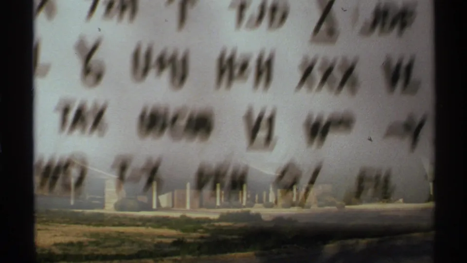 Coded writing remains blurred in the foreground while a low building and open ground appear briefly beyond it in the New Mexico 8mm footage.