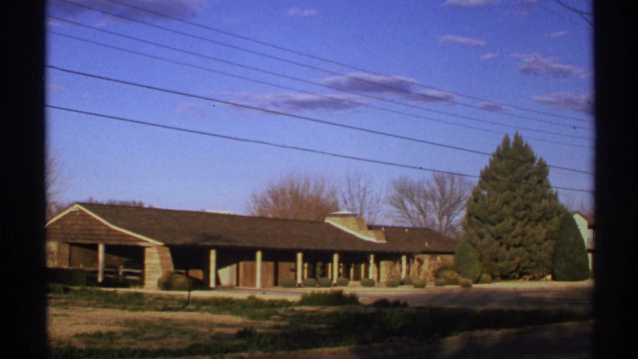 Brown-roofed building, covered walkway, evergreen tree, overhead wires, and blue sky with clouds.