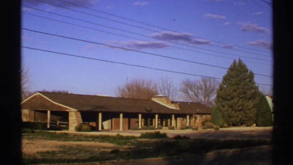8mm home-movie frame from 1972 New Mexico showing a low exterior building, utility wires, bare trees, and a prominent evergreen.