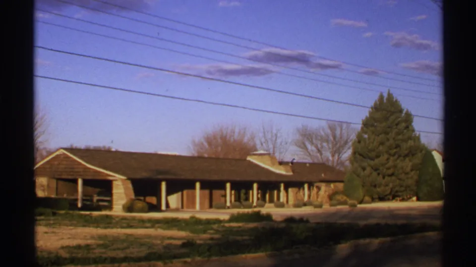 Final 1972 New Mexico frame of the quiet building exterior, evergreen tree, overhead wires, and blue sky with light clouds.