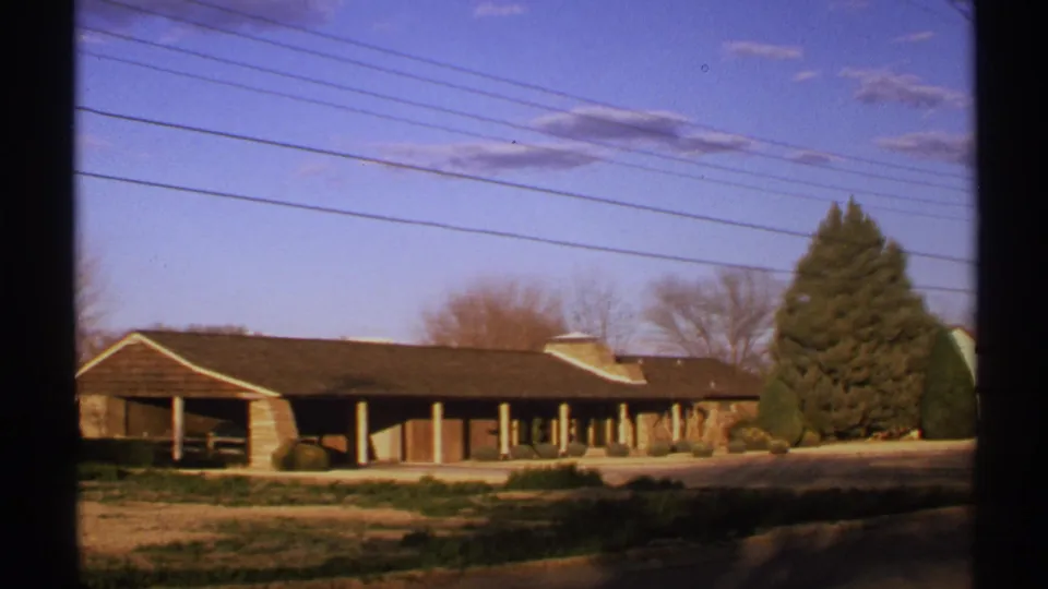 1972 New Mexico 8mm frame showing a low adobe-style building, open roadside ground, bare trees, and overhead power lines.