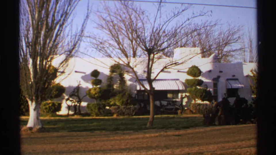 1972 New Mexico home-movie frame showing a white adobe-style building partly screened by winter trees and shaped landscaping.