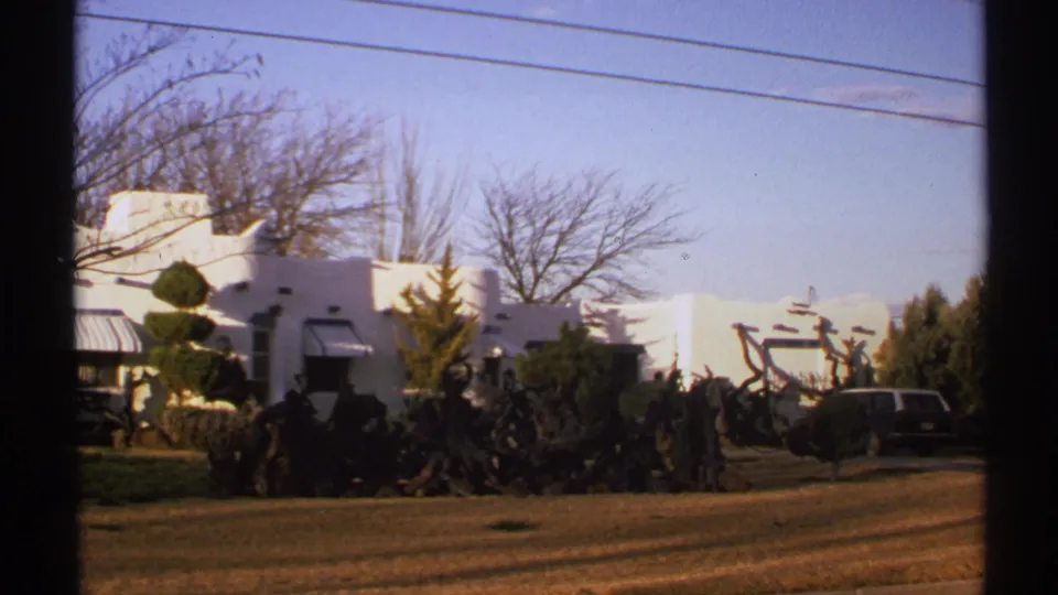 1972 New Mexico 8mm frame showing a white adobe-style exterior, overhead wires, roadside ground, and a parked station wagon at the edge.