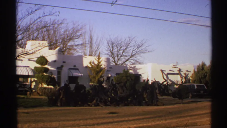 1972 New Mexico 8mm frame showing a white adobe-style building behind manicured shrubs, winter trees, overhead lines, lawn, and a parked vehicle.