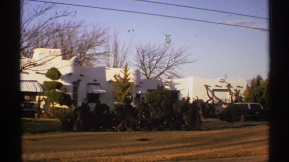 Soft 8mm frame from New Mexico in 1972 showing a white adobe-style exterior, manicured plantings, dry lawn, bare trees, and overhead utility lines.