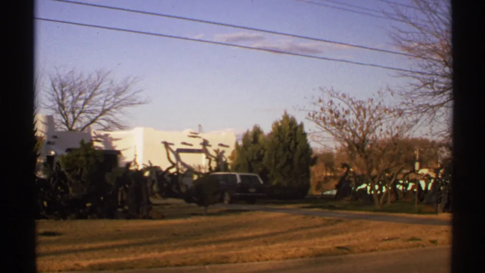 1972 New Mexico 8mm view with a white adobe-style building, dark station wagon, shrubs, winter trees, curbside lawn, and wires under a clear sky.