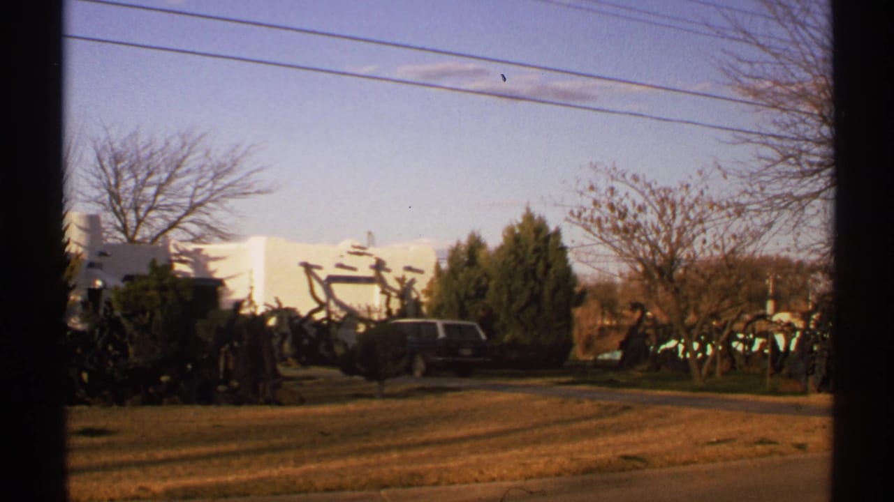 White stucco building and dark station wagon seen past a window frame, with dry grass and bare trees.