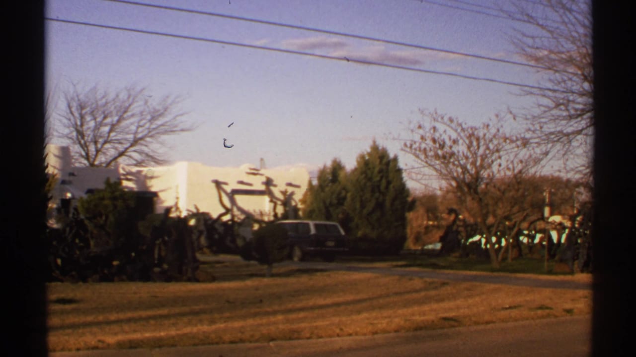 1972 New Mexico exterior with a parked dark station wagon, white stucco structure, dry lawn, bare trees, and abstract metal forms near the entrance area.