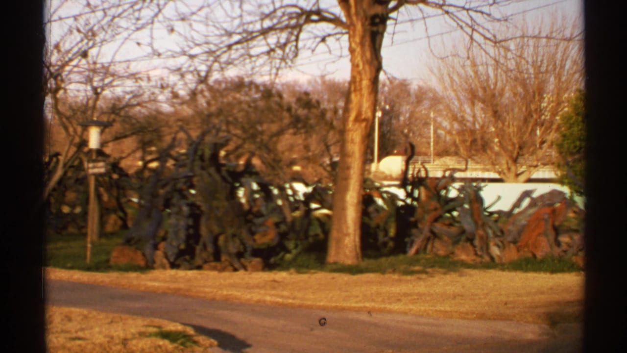 1972 New Mexico home-movie frame showing a roadside outdoor area with bare trees, dry grass, curved pavement, and a large dark abstract sculpture.