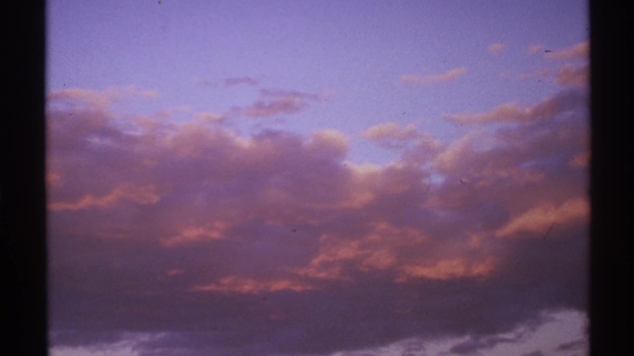 A New Mexico travel-film frame from 1972 shows layered sunset clouds with magenta highlights and a heavy dark frame edge.