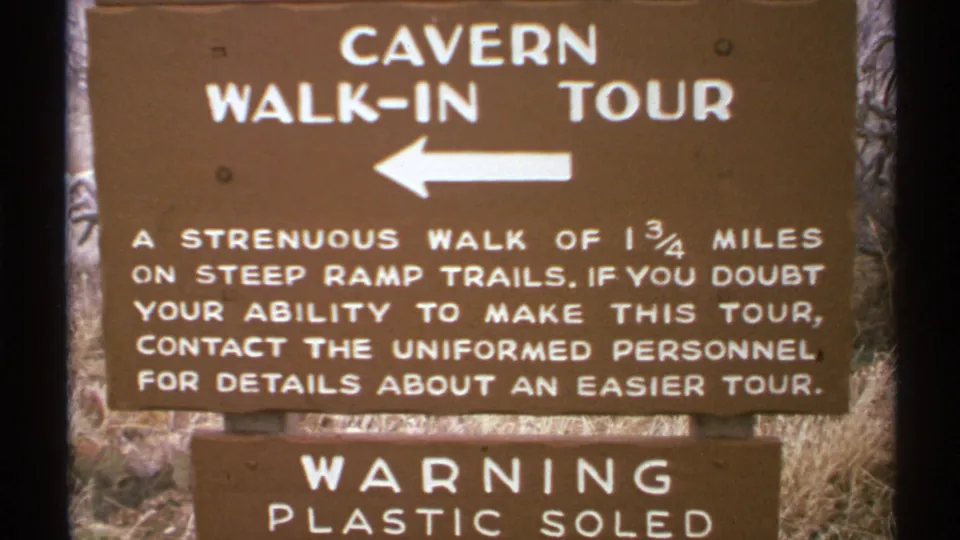Close 8mm view of the New Mexico cavern walk-in tour sign, showing the left arrow, strenuous-walk message, and partial plastic-soled warning.