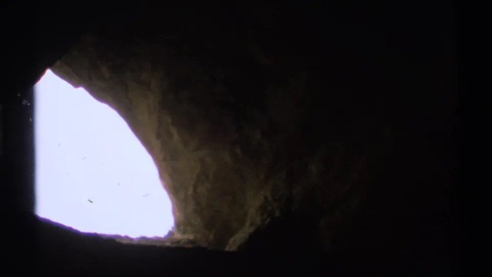 The camera shifts within the cave, keeping the bright exterior opening at left while rough interior rock fills the shadowed frame.