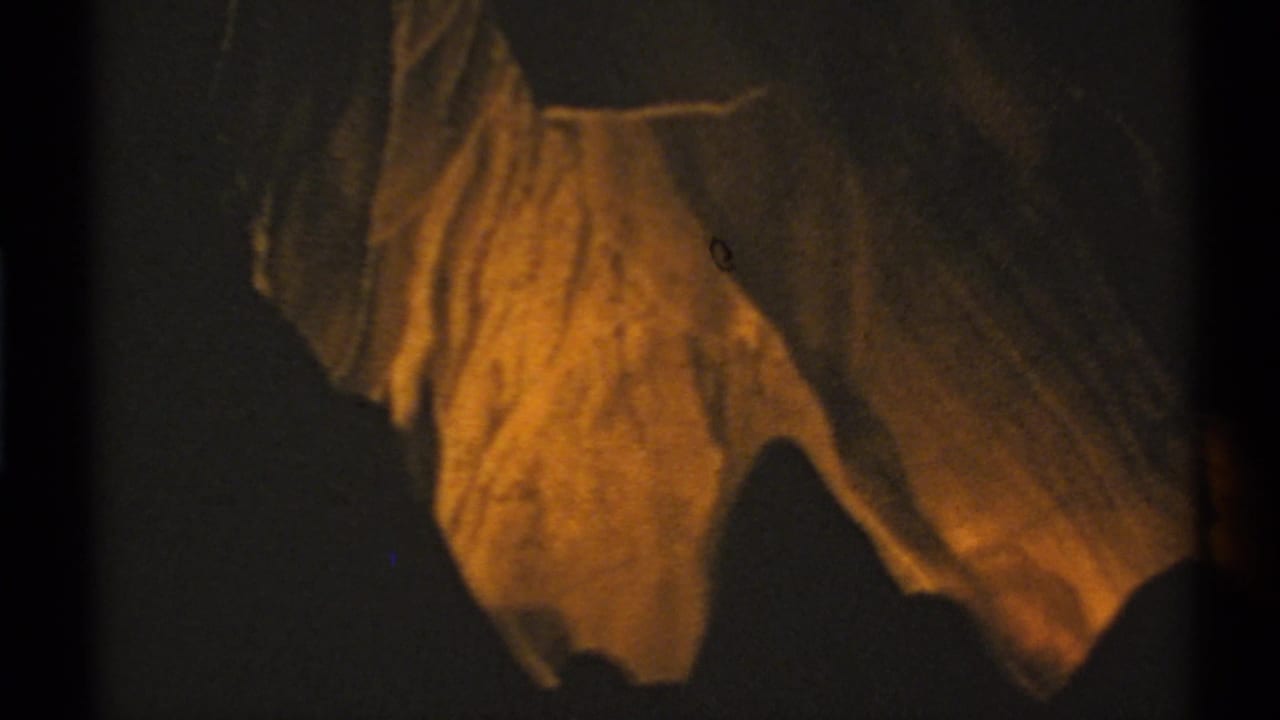 1972 New Mexico 8mm frame showing amber-lit hanging rock forms inside a dark cave interior.