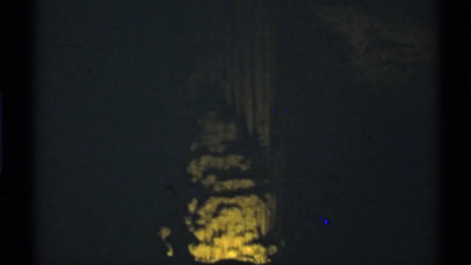 Low-light 8mm frame from a New Mexico cave, showing a small illuminated rock formation below a dark wall.