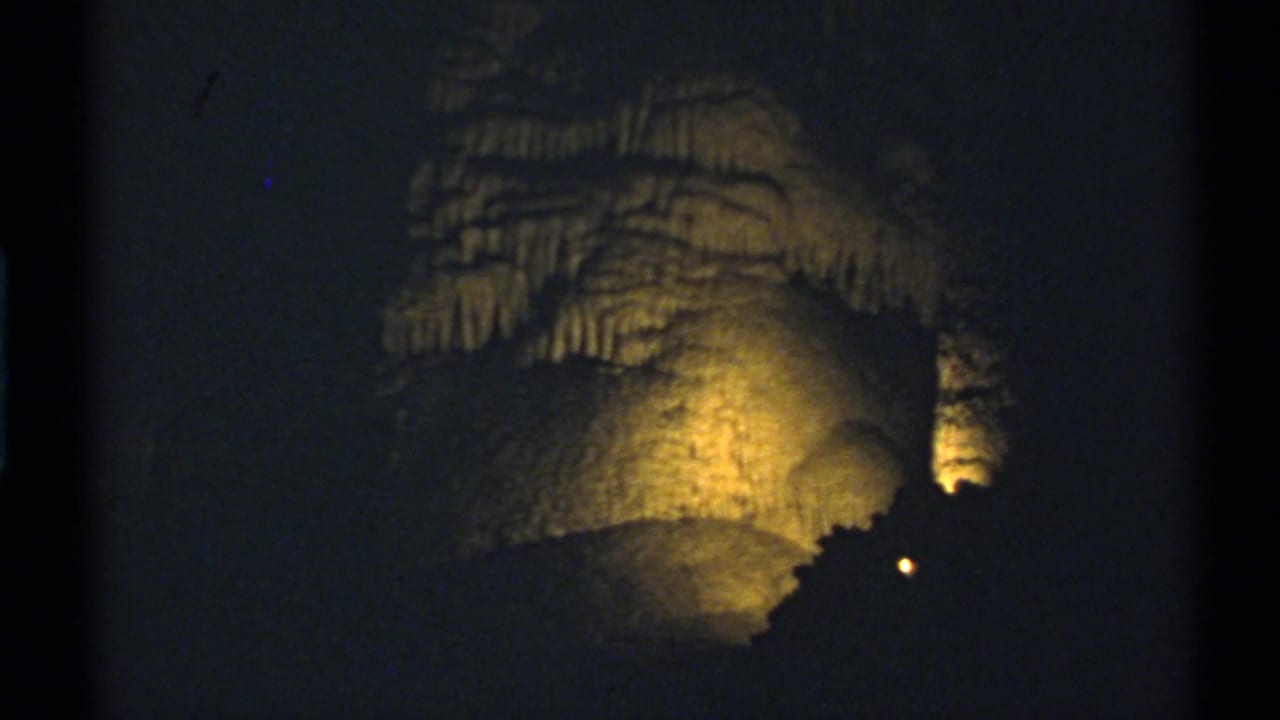 1972 8mm New Mexico cave interior with warm illumination on layered rock walls, stalactites, and a shadowed foreground formation.