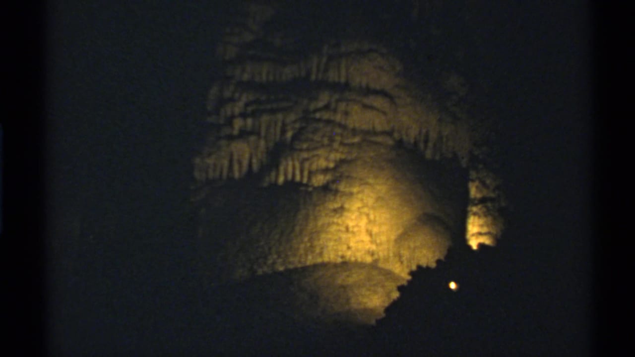 New Mexico cave-tour frame from 1972 showing warm light across a high rock wall, stalactites, and a small point of light in shadow.