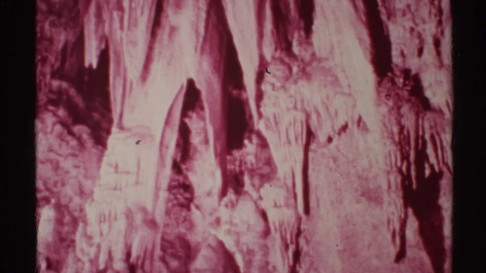 1972 8mm New Mexico cave interior with pink-tinted stalactites, stalagmites, and dark mineral recesses.