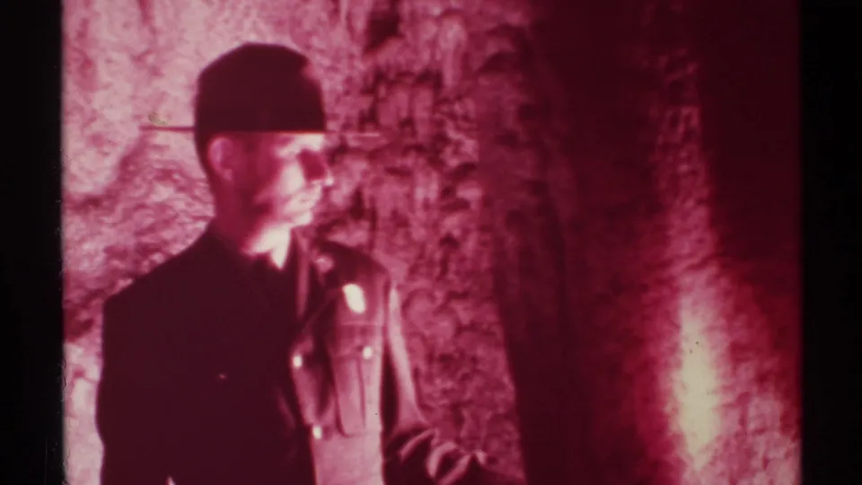 Adult man in a dark cap and uniform-like shirt stands inside a red-lit New Mexico cave, partly silhouetted by exposure.